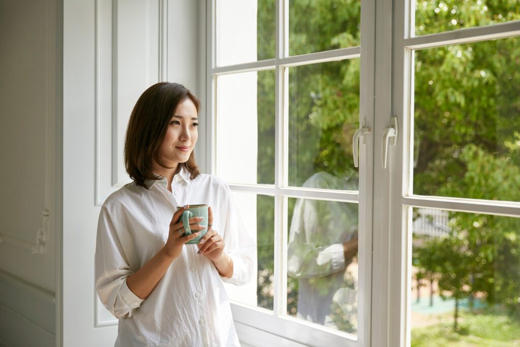 A young woman standing by window at home holding a cup of coffee looking happy and content because of treatment she received for Obsessive-Compulsive Disorder.