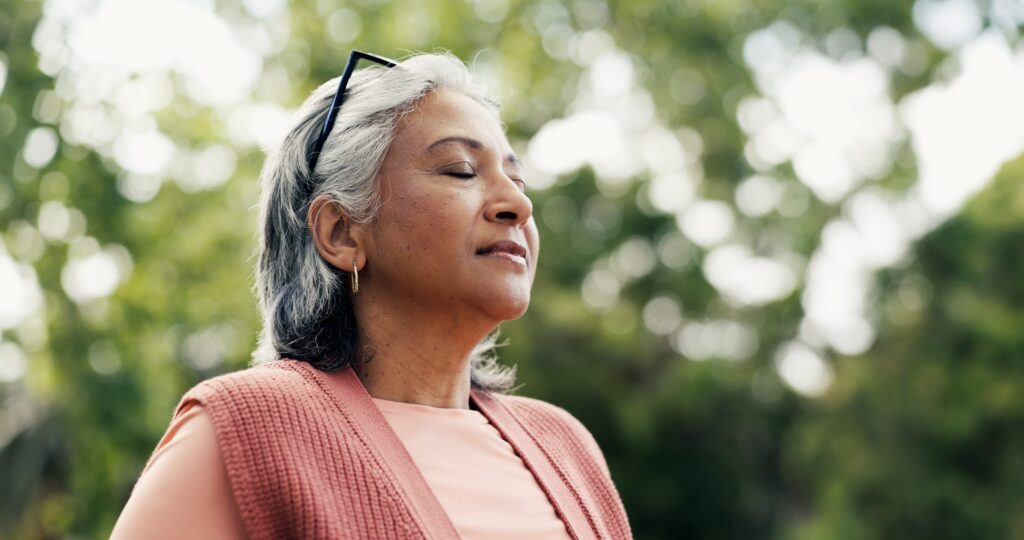 A woman with gray hair holds a deep breath, surrounded by trees.